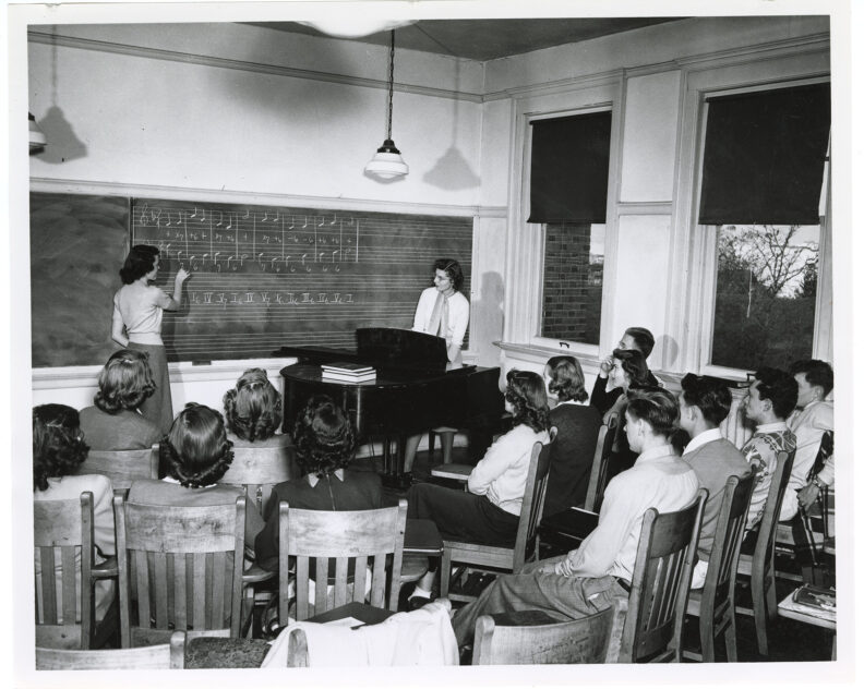 Student writing music on a chalk board in classroom while professor and other students sitting in chairs look on, possibly in the 1950s