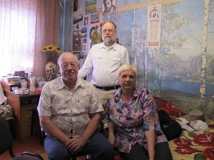 Paul Brians standing behind Lyubov and Victor, in their home with photos and large image of birch tree forest in the background. 