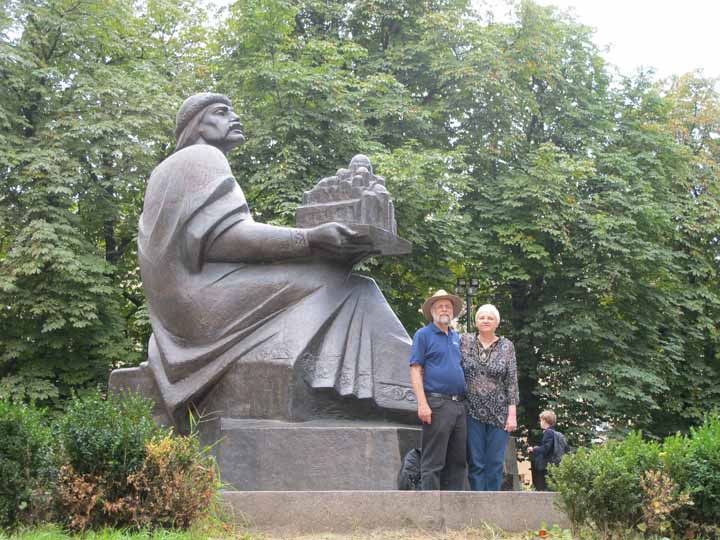 Paul and Lyubov standing by a large statue of a man holding a model of an ornate building. 