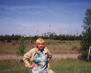 Lyubov standing in front of path and somewhat barren grassy area with trees and station in the background. 