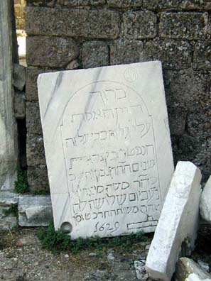 Rectangular white stone tablet with lettering carved into it, lying on ground next to a stone wall.