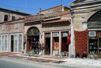 Stone storefronts with colorful rugs and receptacles displayed.