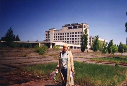 Person standing in front of tall abandoned building with many windows. Parking lot in front of it is overgrown with weeds and trees. 