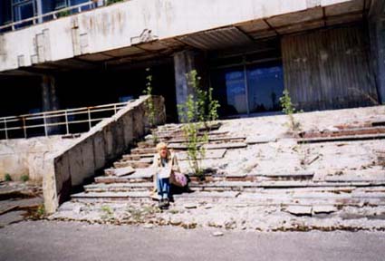Person sitting on crumbling steps in front of an abandoned cement building. Plants are beginning to grow out of the steps. 