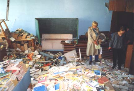 Two people looking at piles of  books and school equipment in an abandoned classroom.
