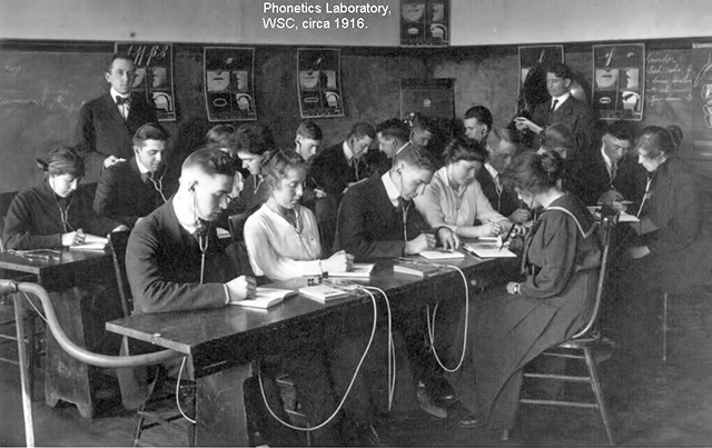 Black & white image of early 20th-century students wearing earphones and writing in notebooks, with 2 male teachers wearing jackets and ties.