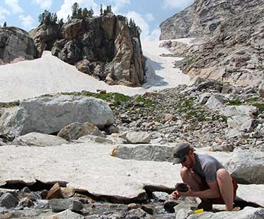 Scott Hotaling squats on a rock at the edge of a glacier and mountain stream.ng. 