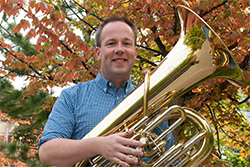 Chris Dickey holding a shiny brass tuba in front of a colorful tree.