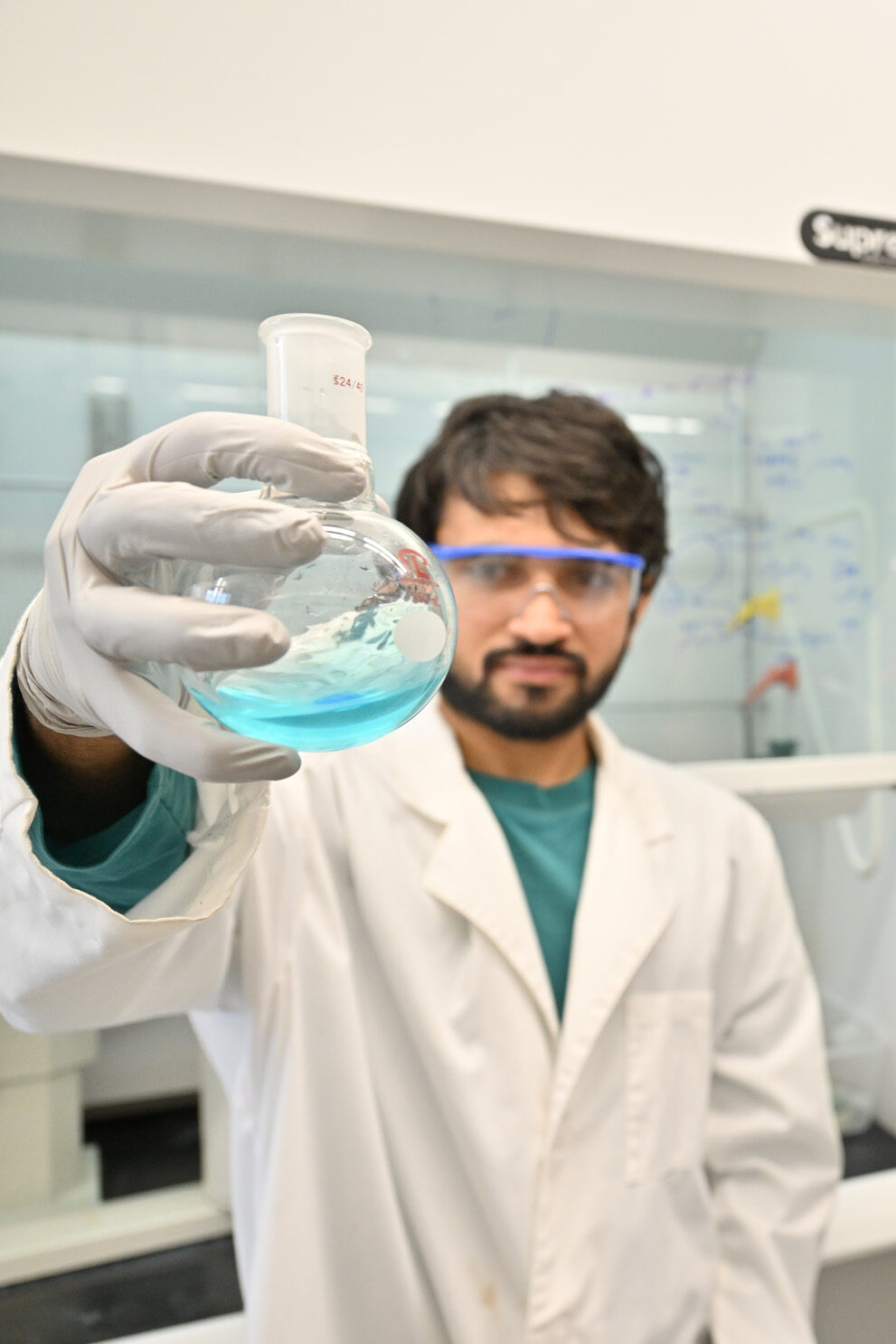 A chemistry student holds a spherical beaker containing a bluish liquid while two other students observe.