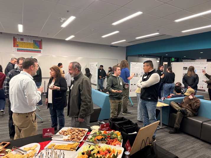 A room full of people conversing. Some research posters are on display, as well as a full table of snacks.