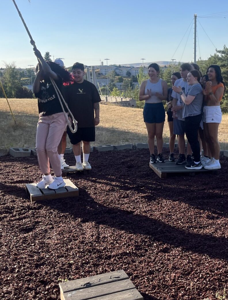 Jo Castaneda cheers on a bridge workshop participant as she jumps to a platform using a rope swing. A group of other bridge participants also look on and cheer.
