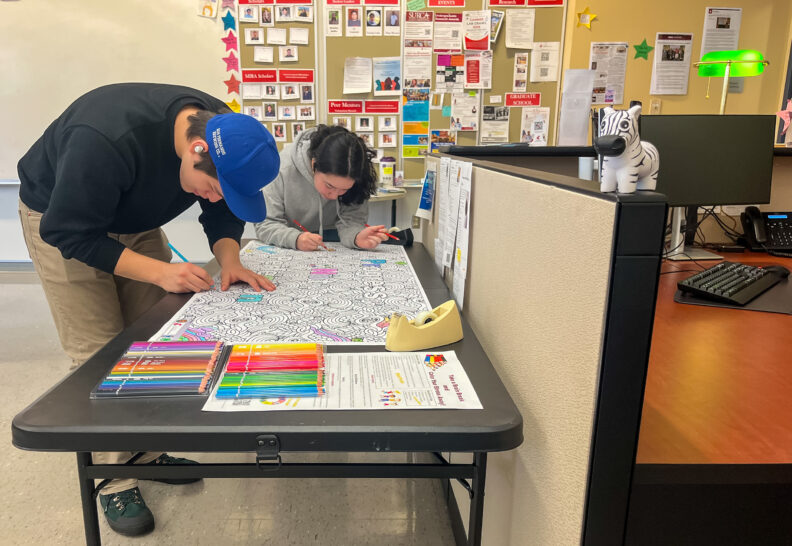 Two STEM students destress at the LSAMP Center by standing over a table and coloring in a motivational poster with color pencils. The poster says “Progress Not Perfection.”
