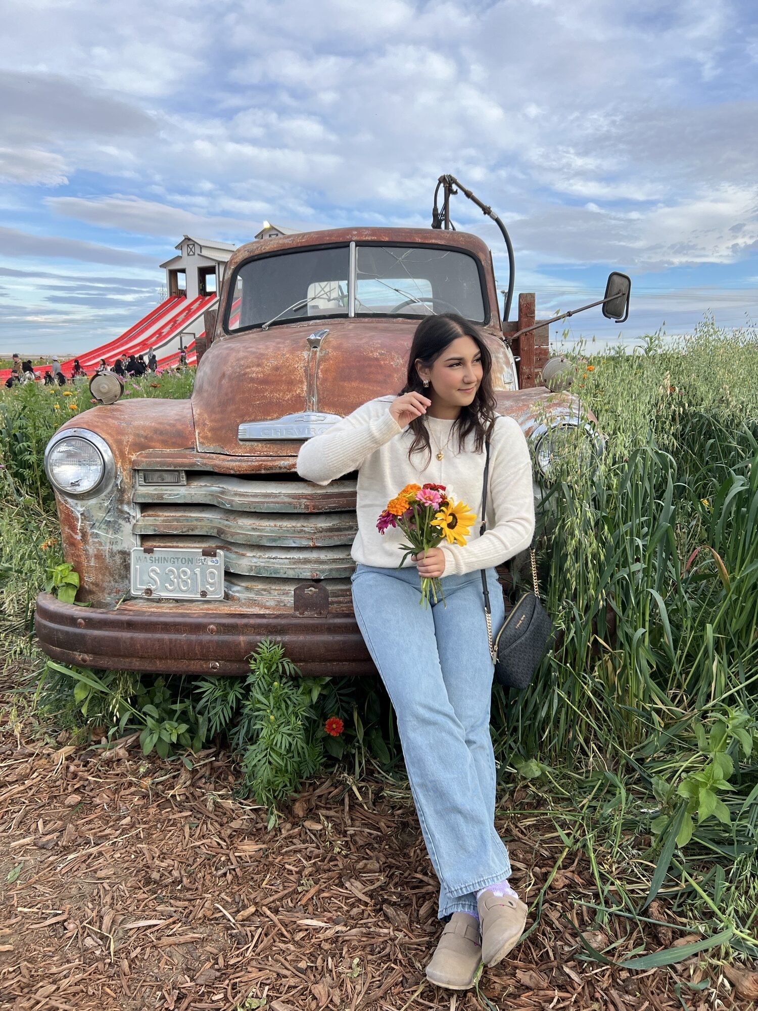 Noemi Barajas holds a bouquet of yellow flowers as she leans against an old pickup truck parked at the edge of a farm field.
