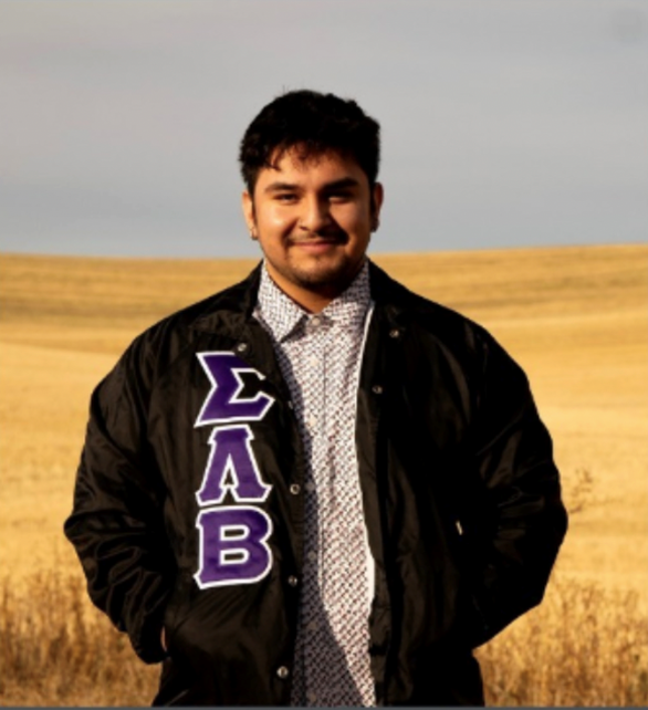 Jo Castaneda stands in front of a wheat field and wears a Sigma Lambda Beta fraternity jacket.