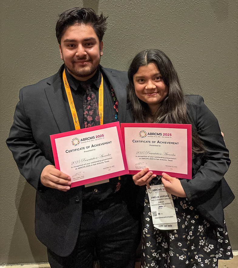 Two professionally dressed students smile as they proudly hold up certificates of achievement. It is seen that the certificates are for winning a presentation award at the ABRCMS 2025 conference.