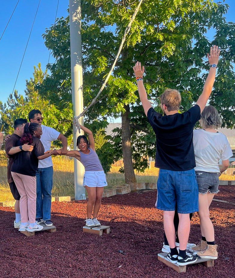 A group of LSAMP students engage in a teambuilding exercise at the University Recreation challenge course. Three students help a fourth student move between small, short wooden platforms without stepping off onto the surrounding ground as three other students look on and cheer from another platform.