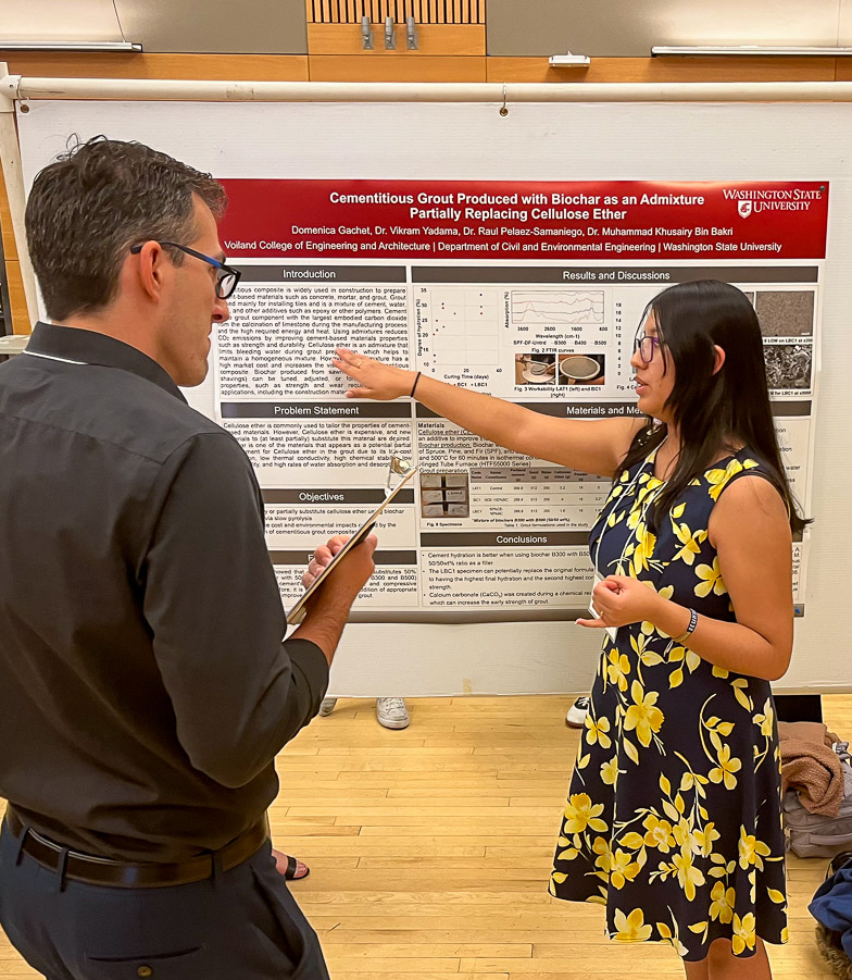 A student presents her poster to a research professional. She points to the abstract section while making a  point as the professional looks on with interest.