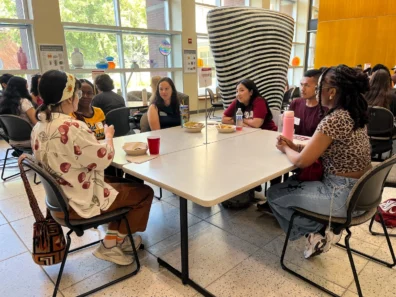 Students attend a professional development session at the Summer Bridge Workshop. They sit around a large square table in the CUE atrium.