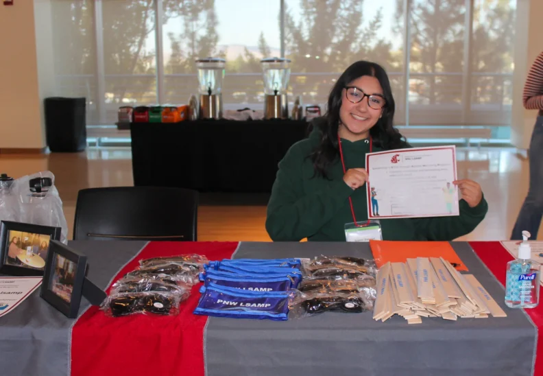 A WSU LSAMP student holds up a certificate while manning a table at a STEM opportunities fair event.