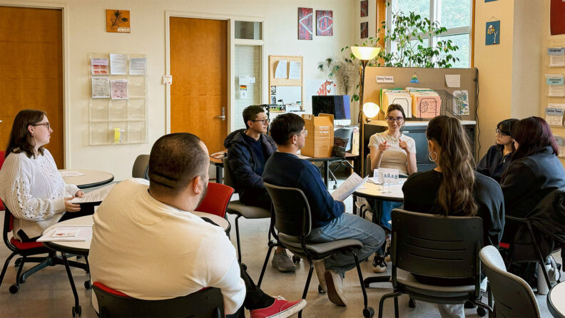 Undergraduate Writing Consultants host a workshop in the writing center. A consultant speaks as undergraduate students seated around a circular table listen.