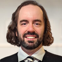 Daniel Rieck, who has shoulder-length, wavy brown hair and a full beard and a black sweater, collared shirt, and tie.