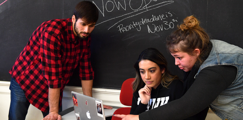 Two students assisting a young woman on her laptop. The students helping are leaning over to look at the laptop and one student is pointing out something on the screen.