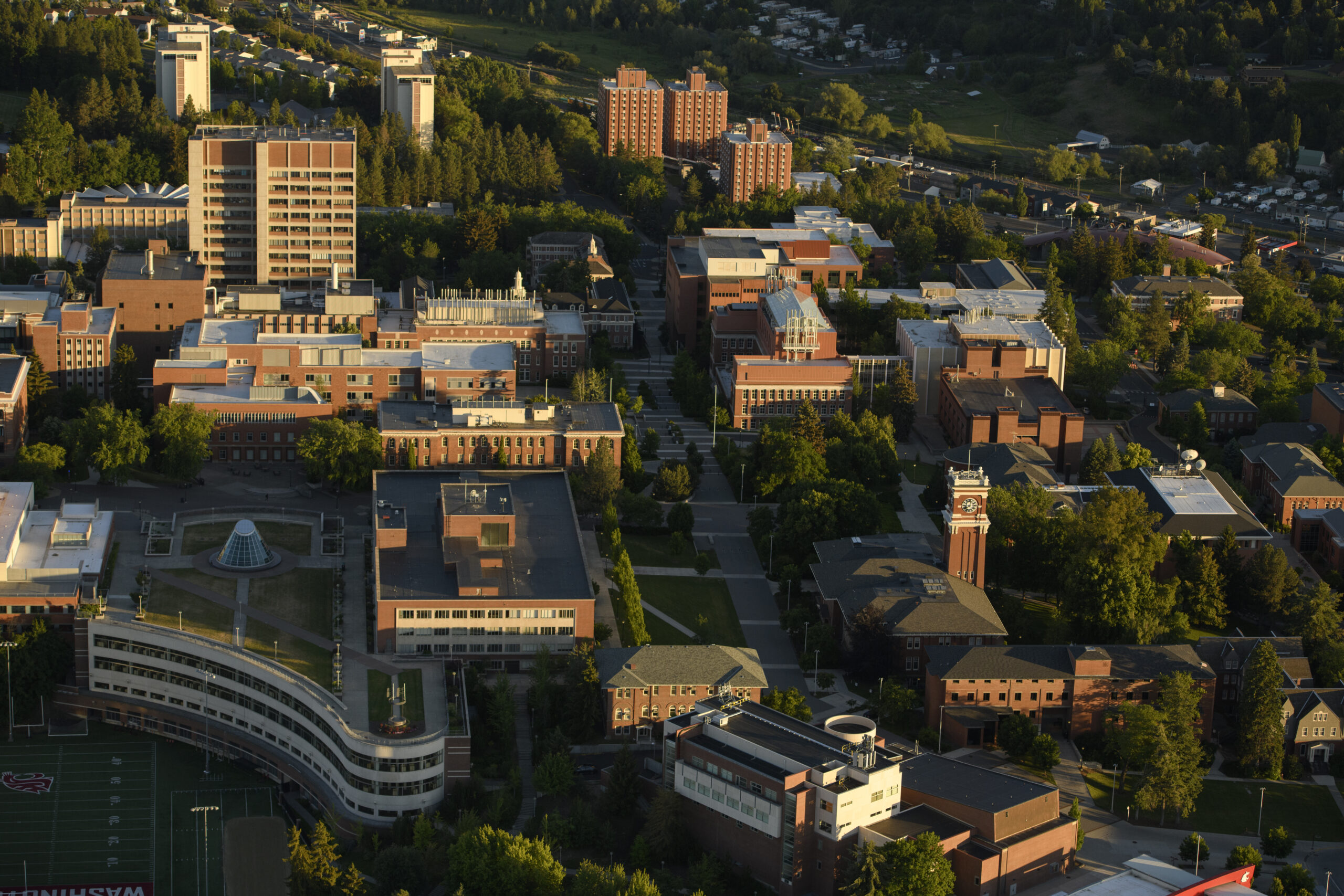 Aerial view of the WSU Pullman campus looking south from high above the lower soccer field near Smith gym. Many prominent campus buildings can be seen and are lit by golden light from the setting sun.