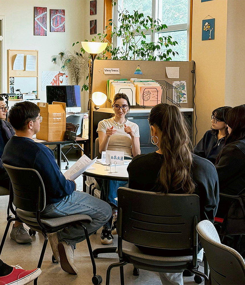 A writing consulting leads a workshop in the Undergraduate Writing Center. Several students are gathered around and listening.
