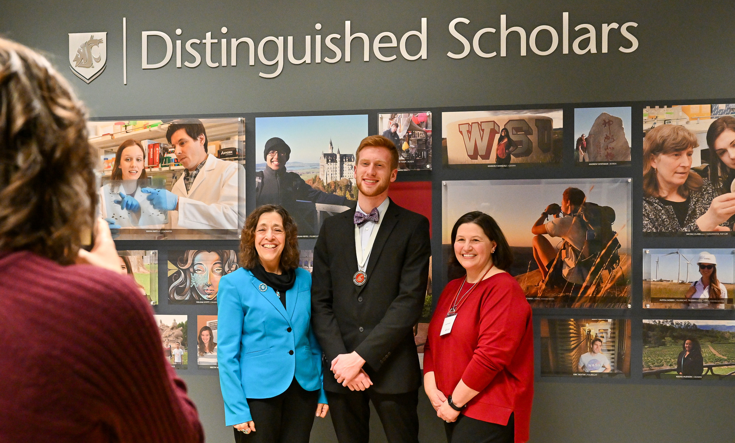 Vice provost Mary Sánchez Lanier, distinguished scholar Jacob Buursma, and director April Seehafer pose for a celebratory photo in front of the Distinguished Scholars gallery wall in the CUB.