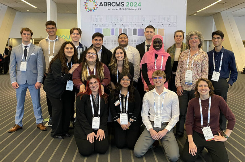 A group of 15 WSU students and faculty members standing and kneeling before a large poster with the ABRCMS conference logo.