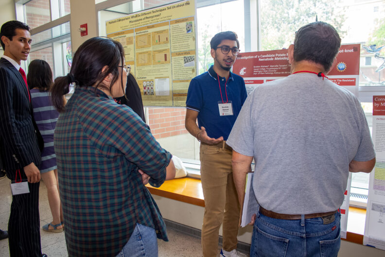 A summer undergraduate researcher explains his research poster to two listening faculty members at the 2024 Summer Research Symposium.