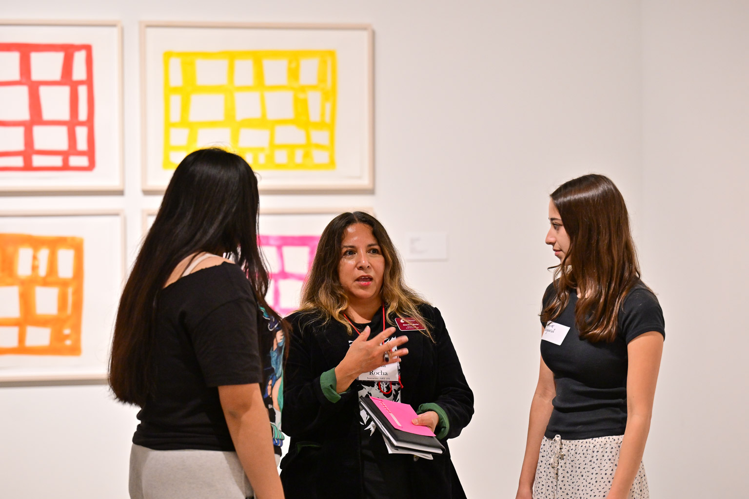 Two students and professor have a conversation at the “Fall 2025 First-Year Programs Evening at the Museum” event. Some colorful paintings are seen behind them.