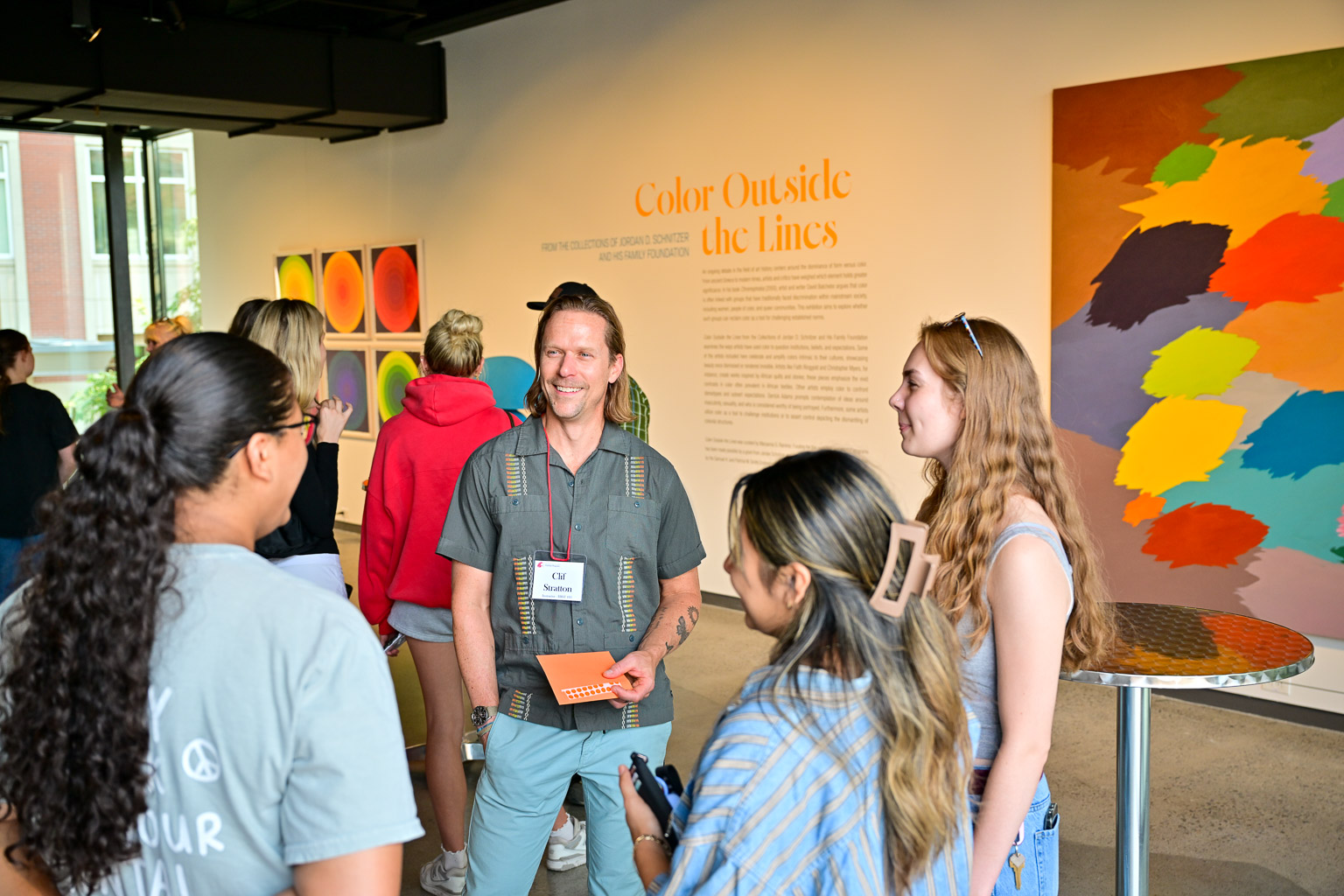 Three students speak with a professor at the “Fall 2025 First-Year Programs Evening at the Museum” event. The introduction to the “Color Outside the Lines” exhibit can be seen on the wall behind them.