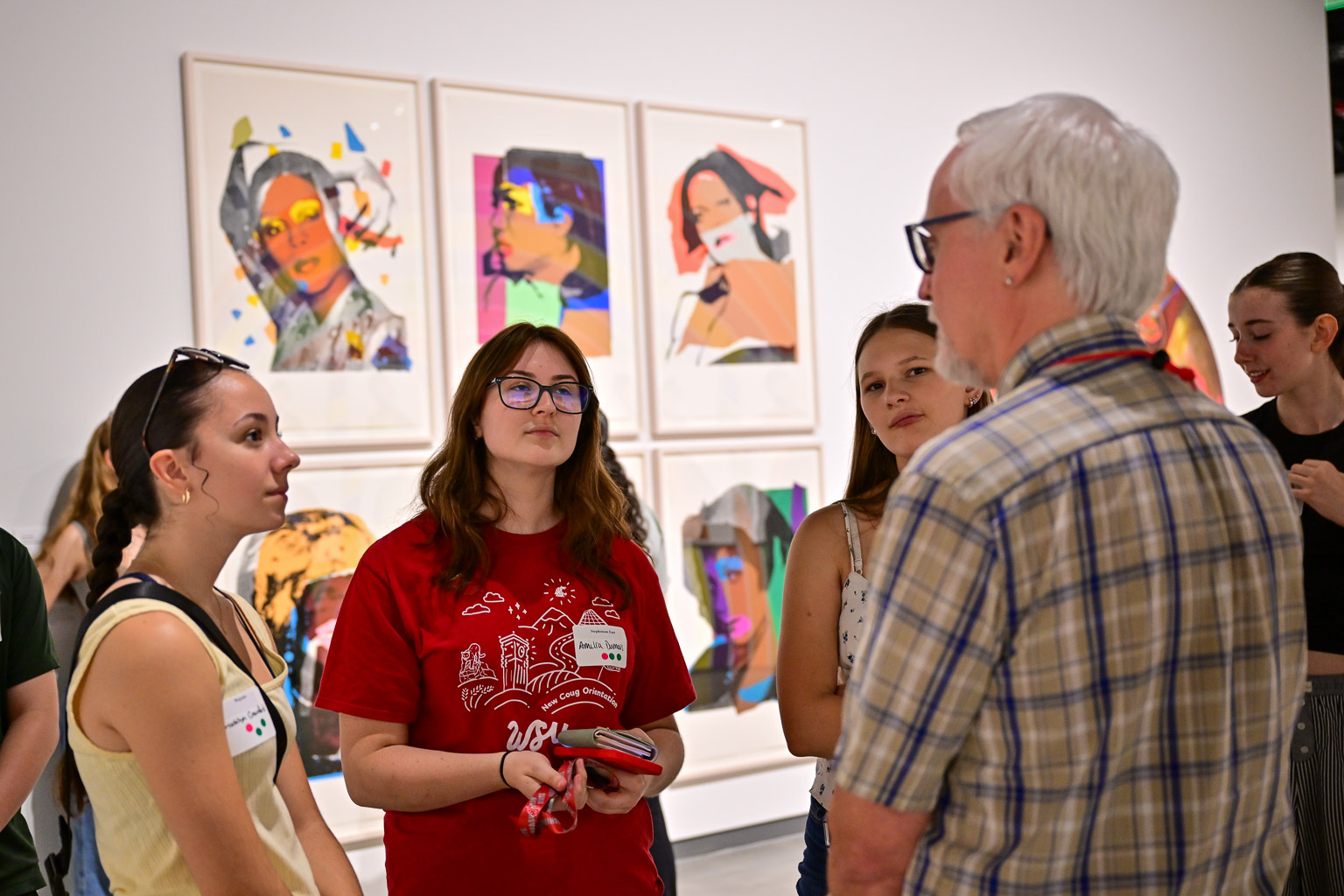 Four students have a conversation with a professor at the “Fall 2025 First-Year Programs Evening at the Museum” event. A grid of photographic art is seen on the wall in the background behind them.