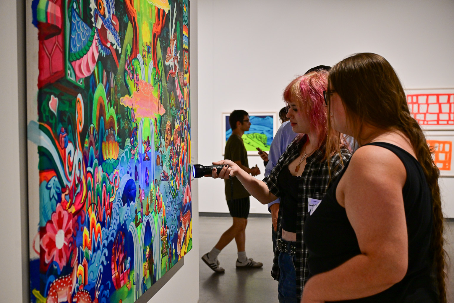 Two students view a vibrant painting at the “Fall 2025 First-Year Programs Evening at the Museum” event. They shine a UV flashlight onto the paintings surface to reveal hidden details.