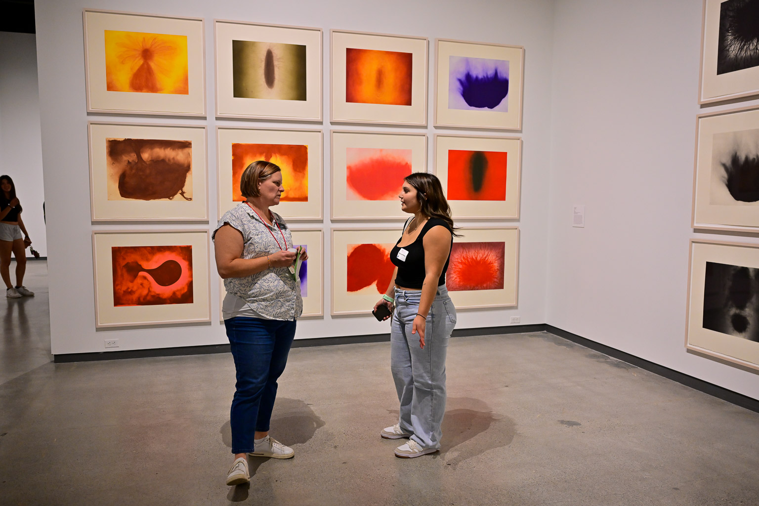 A student and professor have a conversation at the “Fall 2025 First-Year Programs Evening at the Museum” event. A grid of colorful paintings is seen behind them.