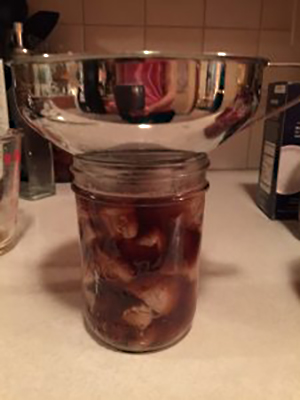 Jar of hot-packed meat positioned under a wide metal canning funnel as boiling broth or liquid is added to cover the meat.