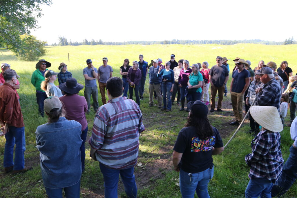 A group outdoors at the edge of a prairie listens to a main speaker present.
