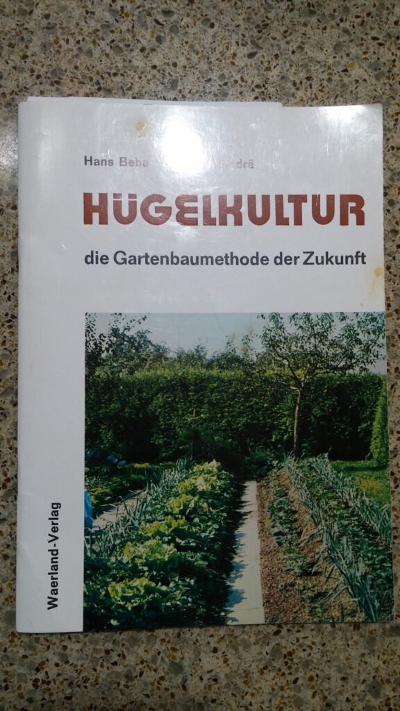 Book cover titled “Hügelkultur: die Gartenbaumethode der Zukunft” by Hans Beba and Heinrich Krämer, showing a photograph of raised garden beds and a pathway between vegetable rows.