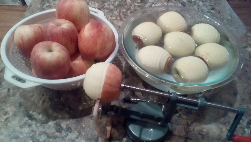 Peeled apples on right in a bowl of water. A colander of unpeeled apples is on the left. Apple peeler in foreground with half-finished apple.