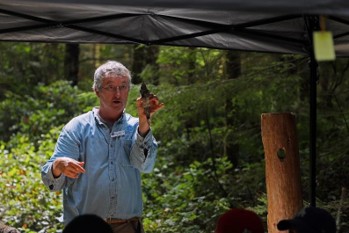 A man stands outside in front of group to give a lecture. He holds up a pinecone.