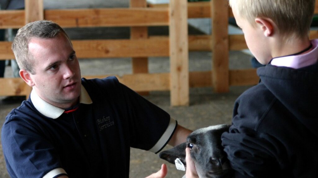 A man speaks to a child who is holding the head of a lamb to show.