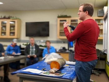 A man stands in a classroom in front of group to give a lecture. He holds up a drill. On the desk in front of him a piece of a log and some papers sit on a blue tarp.