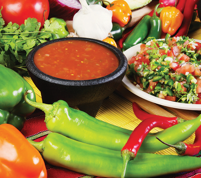 Bowl of red salsa surrounded by fresh peppers, tomatoes, cilantro, onions, and pico de gallo on a table.