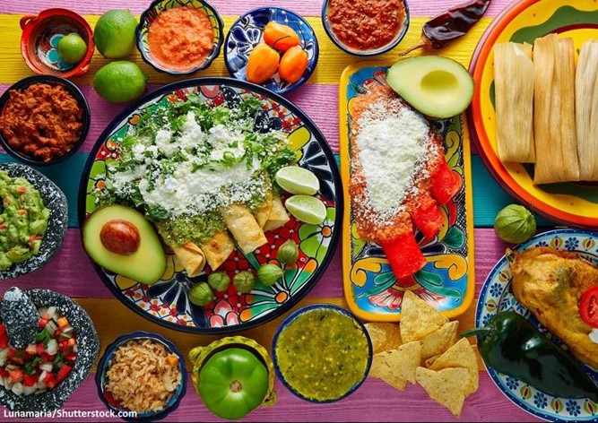 A table spread of Mexican cuisine. The table includes tamales, enchiladas, flautas, green peppers, limes, tortilla chips, rice, guacamole, pico de gallo, beans, and avocados.