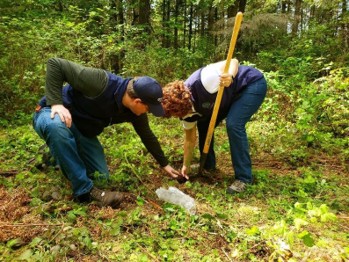 A woman holds a shovel and crouches over a small hole in the forest. A man kneels over and moves dirt near the same hole.