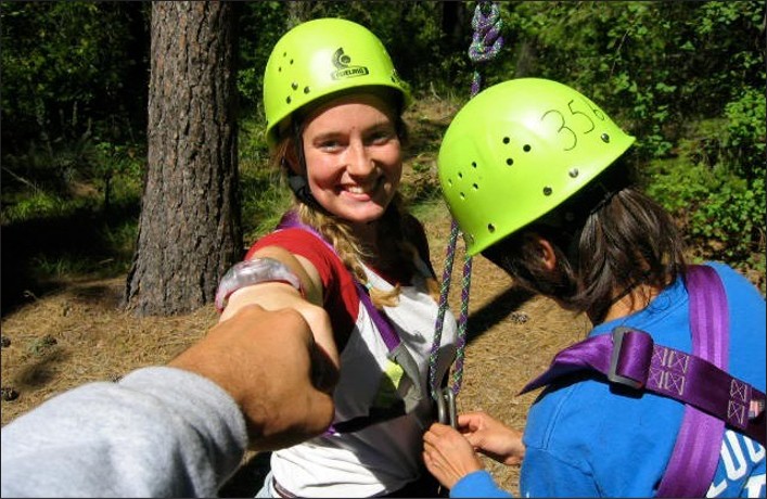 A girl ready for a climbing exercise fist bumps the picture-taker while a second climber adjusts her harness.