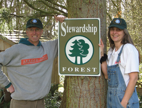 Man and woman standing by a pine tree with a sign that says Stewardship Forest.