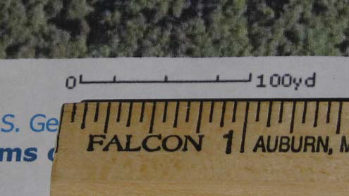 Close-up of a wooden ruler aligned with a map’s printed scale labeled 0–100 yd, showing a measurement comparison.