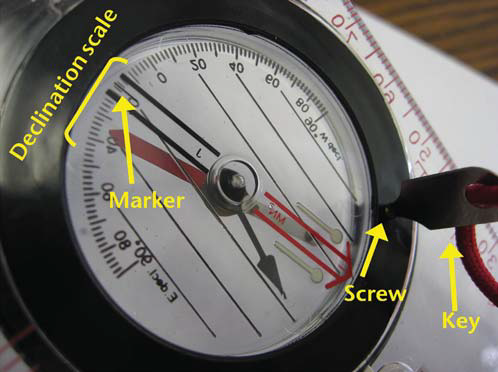 Close-up of a compass bezel with yellow callouts labeling the declination scale, the arrow marker, and the adjustment screw with key inserted.